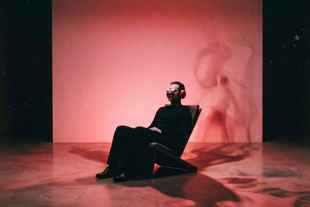 Hombre sentado en una silla con medias pelotas de ping pong sobre los ojos y auriculares, iluminado en rojo, recreando el protocolo del experimento Ganzfeld de privacion sensorial